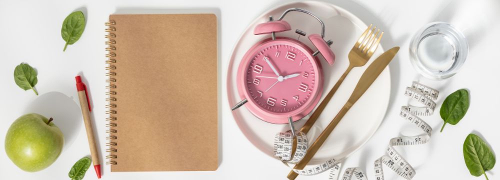 notebook, apple, water ,pen, utensils, measuring tape and pink clock scattered on white table